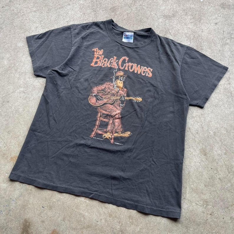 May include: Dark gray t-shirt featuring the text "The Black Crowes" in orange. The graphic depicts a cartoon figure playing a guitar while seated on a stool. The figure is wearing a suit and hat.