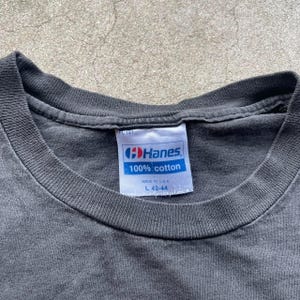 May include: Close-up of a dark gray t-shirt with a white tag. The tag reads "Hanes 100% cotton" and includes the size "L 42-44". The shirt's collar is visible, and the fabric appears worn.