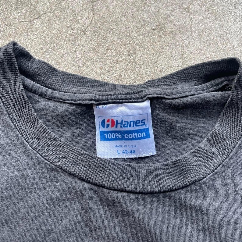 May include: Close-up of a dark gray t-shirt with a white tag. The tag reads "Hanes 100% cotton" and includes the size "L 42-44". The shirt's collar is visible, and the fabric appears worn.