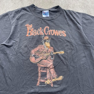 May include: Dark gray t-shirt featuring the band name "The Black Crowes" in orange lettering. The graphic depicts a cartoon crow playing a guitar while seated. The shirt is a Hanes brand, likely vintage.