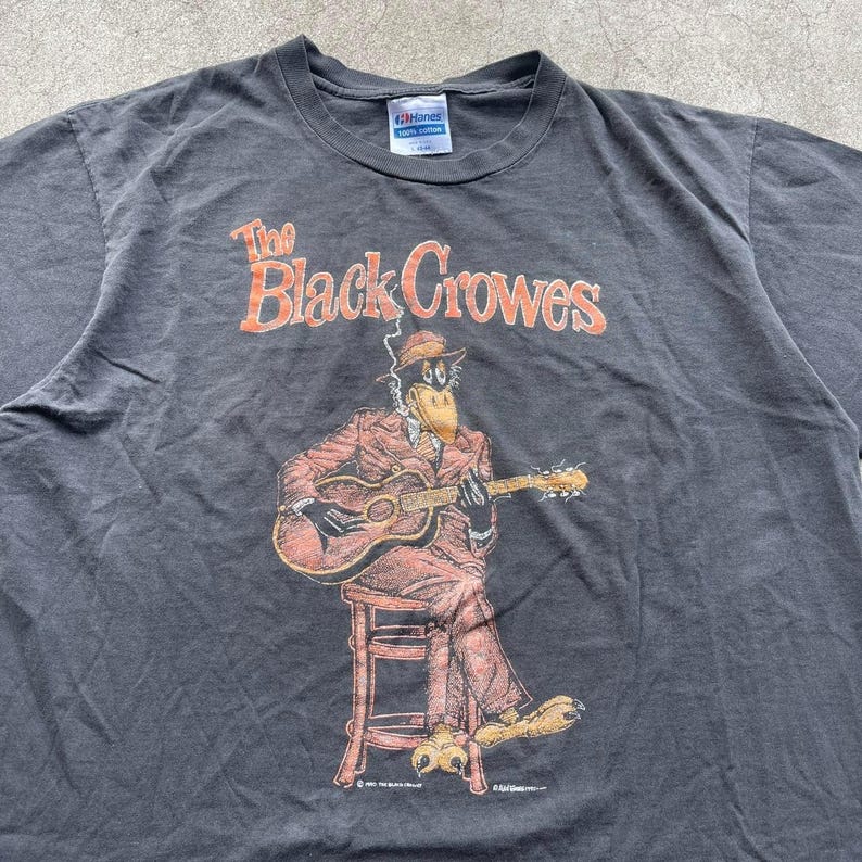 May include: Dark gray t-shirt featuring the band name "The Black Crowes" in orange lettering. The graphic depicts a cartoon crow playing a guitar while seated. The shirt is a Hanes brand, likely vintage.