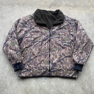 May include: A reversible camouflage jacket with a full zipper closure. The jacket features a brown, green, and tan camouflage pattern on one side and a solid black fleece lining on the other. The jacket has a black collar and cuffs.