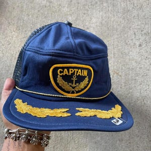 May include: A navy blue mesh trucker hat with a gold embroidered anchor and the word "CAPTAIN". The hat has a gold rope trim and gold embroidered laurel wreaths on the brim.