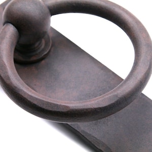 May include: A rustic, dark brown, metal door pull with a ring handle. The handle is attached to a rectangular plate with two visible screws.