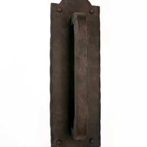 May include: A dark brown, wrought iron door pull with a decorative, arched top and a hammered finish. The pull is mounted on a rectangular plate with a hammered finish.