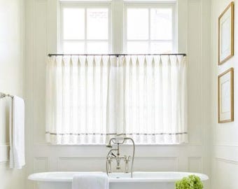 May include: A pair of off-white cafe curtains with vertical pleats hanging from a dark metal rod over a white clawfoot bathtub. The curtains feature a subtle horizontal band near the bottom hem and are positioned in front of a window.