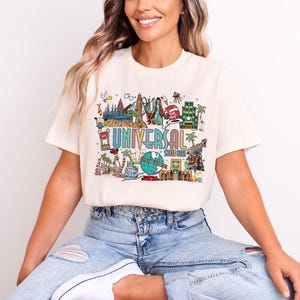 Może przedstawiać: Kremowa koszulka z kolorową grafiką przedstawiającą atrakcje Universal Studios. Projekt zawiera napis "UNIVERSAL STUDIOS" oraz różne elementy parku rozrywki, takie jak globus, kolejki górskie i palmy.