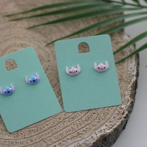 Peut inclure: Deux paires de boucles d'oreilles à clous représentant le personnage Stitch du film d'animation Lilo & Stitch. Une paire est bleue, l'autre rose. Les boucles d'oreilles sont présentées sur des cartes à boucles d'oreilles bleu clair avec un trou en haut, posées sur une surface en bois.