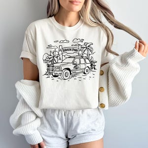 May include: A white t-shirt with a black and white illustration of a Jeep driving through a Jurassic Park gate. The text "Jurassic Park" is visible on the gate.