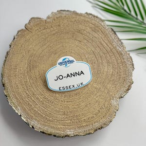 May include: A white name tag with blue trim and the text "Universal Orlando Resort" on top, "Jo-Anna" in the center, and "Essex.uk" on the bottom. The name tag is sitting on a wooden slice.