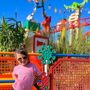 May include: A young girl wearing pink sunglasses and a pink t-shirt with the word "Blinky" on it stands in front of a colorful playground structure. The structure is made of large, colorful blocks and has a green, star-shaped toy on top.
