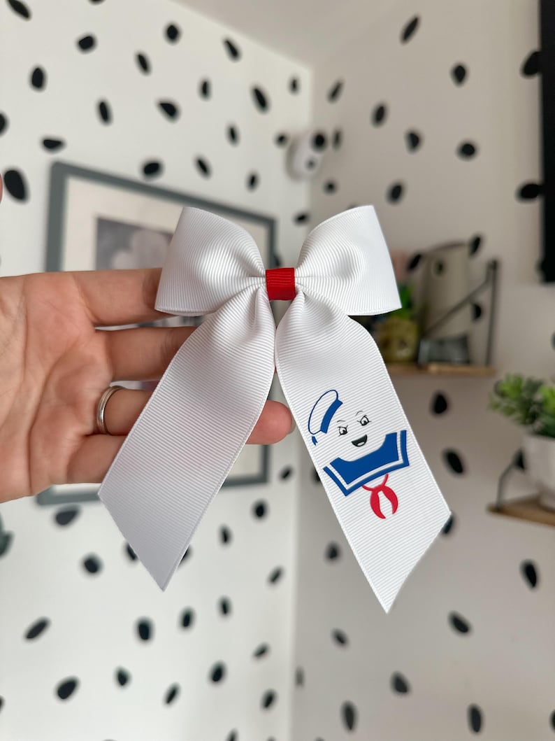 Puede incluir: Lazo blanco con un centro rojo y una cara de fantasma de dibujos animados con un sombrero de marinero azul y una bufanda roja. El fantasma lleva una camisa de marinero azul y blanca a rayas.