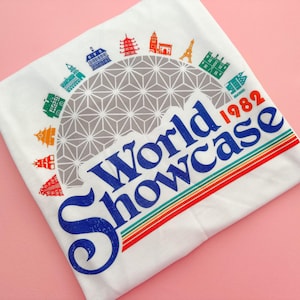 May include: White t-shirt with a graphic design that reads "World Showcase 1982." The design includes a geometric dome, colourful stripes, and illustrations of landmarks. The t-shirt is folded on a pink surface.
