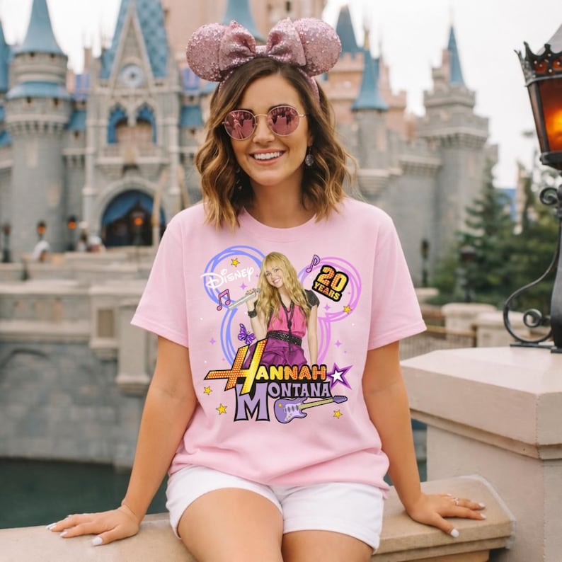 K&ouml;nnte beinhalten: Ein hellrosa T-Shirt mit einer Grafik von Hannah Montana, dem Text "Hannah Montana" und "20 Years". Das Shirt hat ein Disney-Logo und Noten. Die Person tr&auml;gt eine rosa Schleife und eine Sonnenbrille.