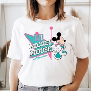 May include: White t-shirt featuring a retro design with the text "The Mickey Mouse" in teal and pink, with a cartoon Mickey Mouse graphic. The shirt also includes "Walt Disney World" text.
