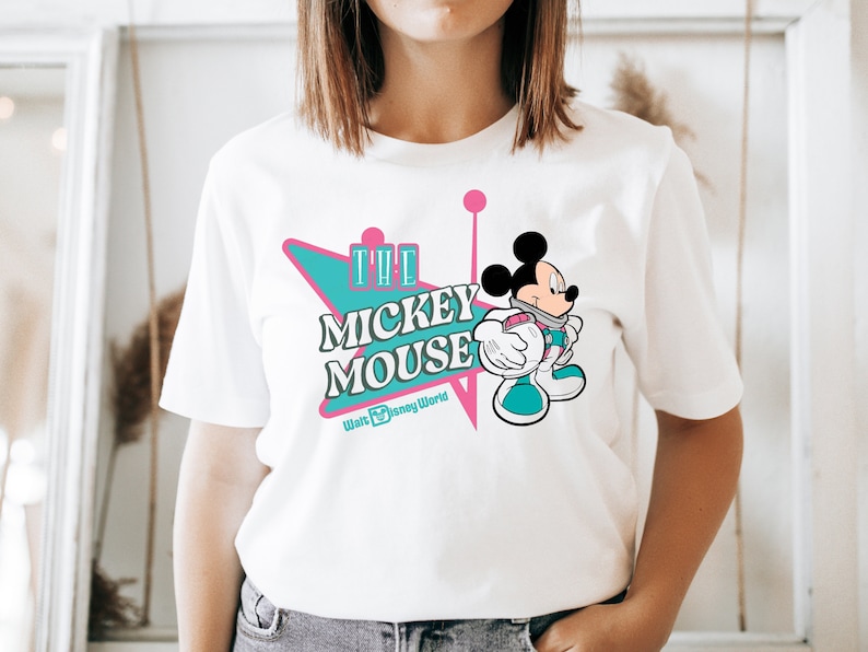 May include: White t-shirt featuring a retro design with the text "The Mickey Mouse" in teal and pink, with a cartoon Mickey Mouse graphic. The shirt also includes "Walt Disney World" text.