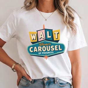 Op de afbeelding: Wit T-shirt met een kleurrijke afbeelding van het Walt Disney's Carousel of Progress-bord. Het ontwerp bevat de tekst "WALT" in gele letters en "CAROUSEL OF PROGRESS" in een turquoise ovaal. Onderaan staat een ster.