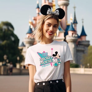 May include: White t-shirt featuring a Mickey Mouse graphic with the text "The Mickey Mouse Walt Disney World". The design includes a retro-style teal and pink graphic. The person is wearing black Mickey Mouse ears.