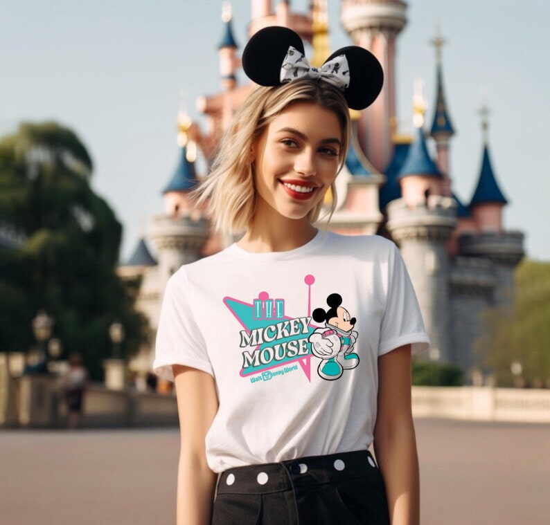 May include: White t-shirt featuring a Mickey Mouse graphic with the text "The Mickey Mouse Walt Disney World". The design includes a retro-style teal and pink graphic. The person is wearing black Mickey Mouse ears.