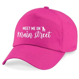 May include: A vibrant pink baseball cap featuring the white text "MEET ME ON main street". The cap has a curved peak and a classic six-panel design, ideal for everyday wear.