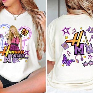 K&ouml;nnte beinhalten: Cremefarbenes T-Shirt mit einem Hannah Montana-Grafik und dem Text "20 Years". Das Frontdesign umfasst eine lila Gitarre, Schmetterlinge und Sterne. Die R&uuml;ckseite hat ein lila "HM"-Logo mit Noten, Sternen und anderen Grafiken.