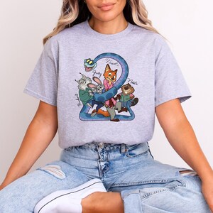 Puede incluir: Camiseta gris claro con un dibujo animado colorido de personajes de una pel&iacute;cula. El dibujo incluye un n&uacute;mero 2 azul, un zorro, un conejo, un castor y un gato. Los nombres de los personajes est&aacute;n escritos encima.