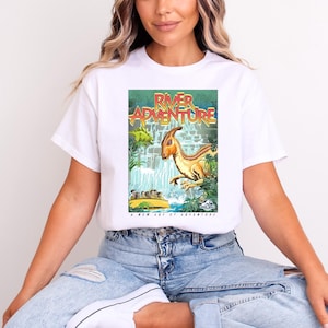 Pode incluir: Camiseta branca com uma estampa gráfica de um dinossauro de desenho animado navegando por um rio em uma jangada. O texto "River Adventure" está impresso em letras amarelas e vermelhas no gráfico. O texto "A New Way of Adventure" está impresso abaixo do gráfico.