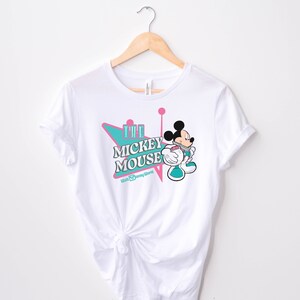 May include: White t-shirt featuring a graphic of Mickey Mouse in a retro style. The design includes the text "The Mickey Mouse" in a teal and pink color scheme, with the Walt Disney World logo below. The shirt is hanging on a wooden hanger.