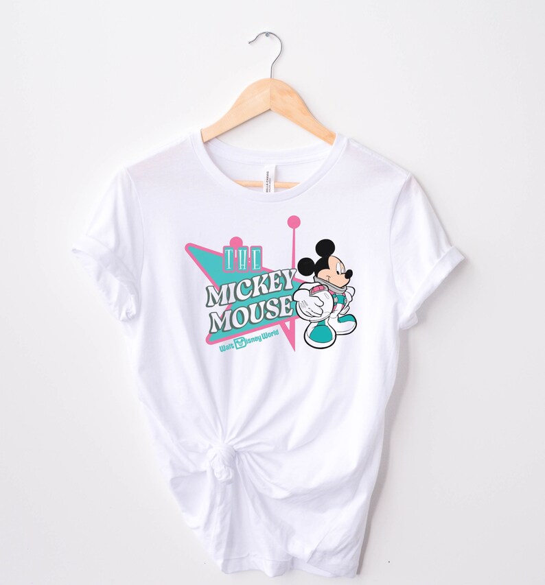 May include: White t-shirt featuring a graphic of Mickey Mouse in a retro style. The design includes the text "The Mickey Mouse" in a teal and pink color scheme, with the Walt Disney World logo below. The shirt is hanging on a wooden hanger.