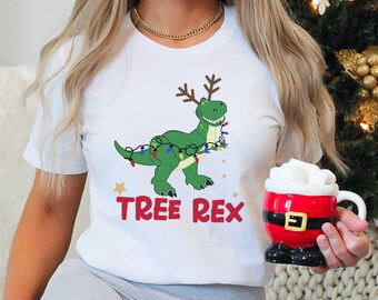 May include: White t-shirt with a green dinosaur wearing antlers and Christmas lights. The text "TREE REX" is printed below the dinosaur. A red and white Santa mug is held in the hand.
