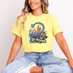 Puede incluir: Camiseta amarilla con un dibujo animado del n&uacute;mero "2" y personajes de animales, incluyendo un zorro, un conejo y un castor. Los nombres "Judy", "Nick" y "Robert" est&aacute;n escritos en la camiseta. La camiseta la lleva una persona.