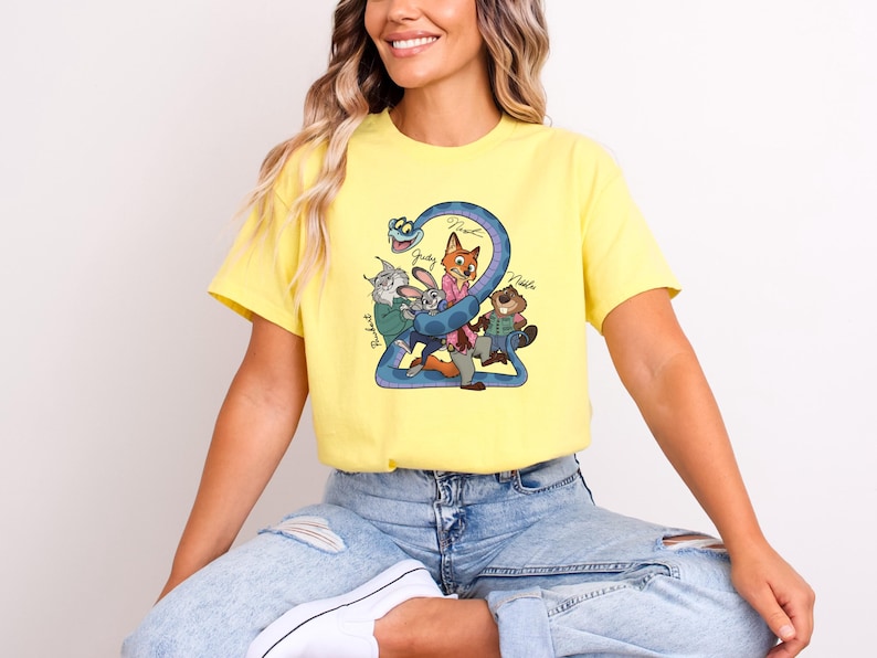 Puede incluir: Camiseta amarilla con un dibujo animado del n&uacute;mero "2" y personajes de animales, incluyendo un zorro, un conejo y un castor. Los nombres "Judy", "Nick" y "Robert" est&aacute;n escritos en la camiseta. La camiseta la lleva una persona.