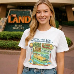 May include: White t-shirt with the text "GET IN LOSER, WE'RE LEARNING ABOUT LETTUCE" and a colorful graphic of a boat ride. The shirt is worn by a person in front of a sign that says "THE LAND WITH NATURE".