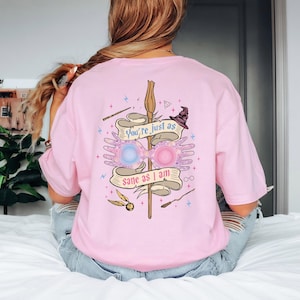 Könnte beinhalten: Hellrosa T-Shirt mit einem Grafikdesign auf der Rückseite. Das Design beinhaltet einen Zauberstab, eine Brille, einen Hexenhut und den Text "You're just as sane as I am". Das Shirt hat eine lockere Passform.