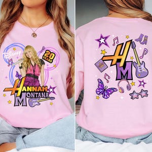 K&ouml;nnte beinhalten: Hellrosa T-Shirt mit einer Grafik von Hannah Montana, dem Text "Hannah Montana" und "20 Years". Das Design umfasst Noten, Sterne und eine Gitarre. Die R&uuml;ckseite des Shirts hat ein passendes Design.
