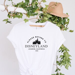 Puede incluir: Camiseta blanca con un gráfico negro del castillo de Disneyland y el texto "Please Return to Disneyland Anaheim, California 1955."