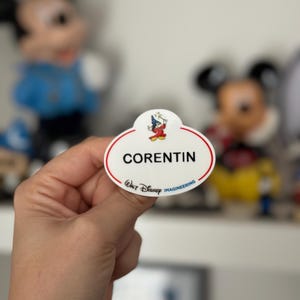 Op de afbeelding: Een wit ovaal naambadge met de naam "Corentin" in zwarte tekst. Het badge heeft een klein cartoonbeeld van Mickey Mouse verkleed als tovenaar. De tekst "Walt Disney Imagineering" is in blauw en zwart onder de naam gedrukt.