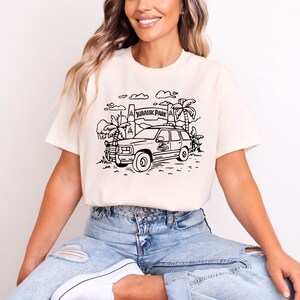 May include: A white t-shirt with a black line drawing of a car driving through a Jurassic Park entrance. The text "Jurassic Park" is on the sign.