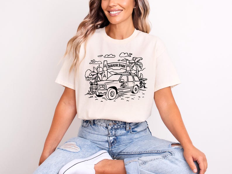 May include: A white t-shirt with a black line drawing of a car driving through a Jurassic Park entrance. The text "Jurassic Park" is on the sign.