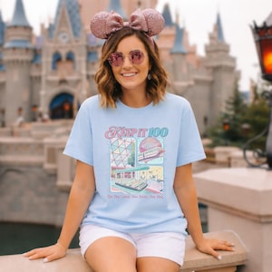 Op de afbeelding: Lichtblauw T-shirt met de tekst "KEEP IT 100" en een kleurrijke afbeelding van een pretpark. De afbeelding bevat afbeeldingen van een geodetische bol, een monorail en een boottocht. De zin "On The Land, The Sea, The Sky" staat eronder.