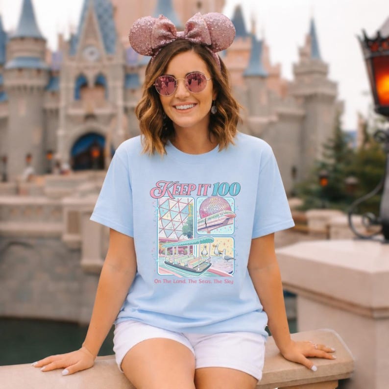 Op de afbeelding: Lichtblauw T-shirt met de tekst "KEEP IT 100" en een kleurrijke afbeelding van een pretpark. De afbeelding bevat afbeeldingen van een geodetische bol, een monorail en een boottocht. De zin "On The Land, The Sea, The Sky" staat eronder.