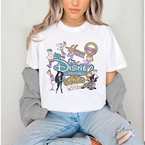 May include: White t-shirt featuring a colorful graphic design with the text "Disney Channel" and cartoon characters. The design includes characters from "Hannah Montana" and "The Suite Life". The shirt is worn by a person.