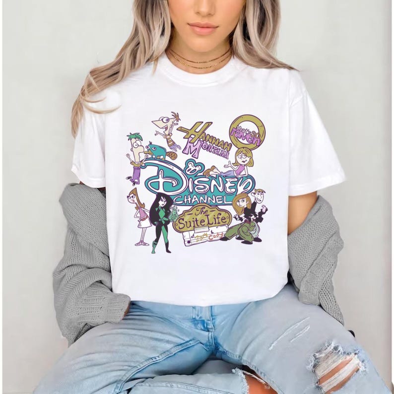 May include: White t-shirt featuring a colorful graphic design with the text "Disney Channel" and cartoon characters. The design includes characters from "Hannah Montana" and "The Suite Life". The shirt is worn by a person.