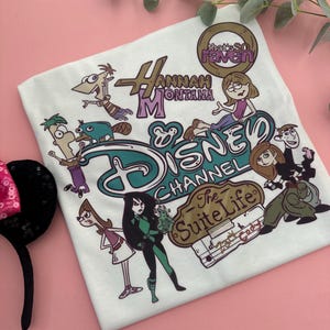 May include: White t-shirt featuring Disney Channel characters from Hannah Montana, That's So Raven, Kim Possible, and The Suite Life of Zack & Cody. The shirt has a teal and brown Disney Channel logo.