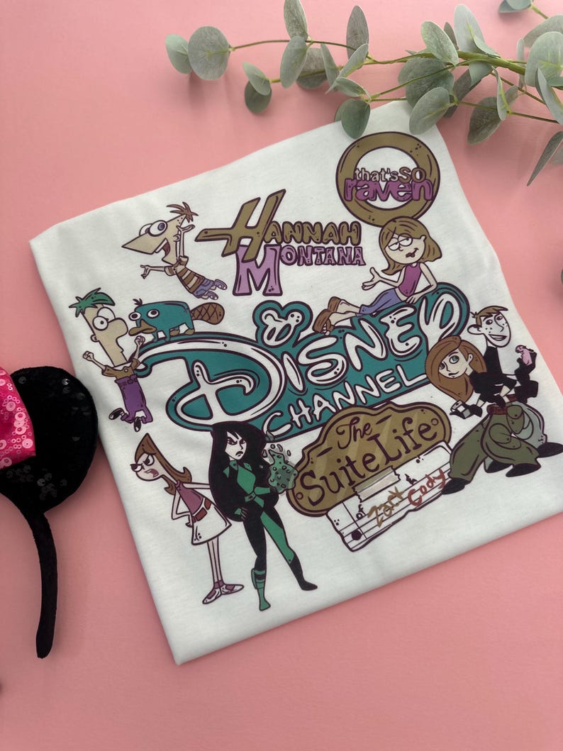 May include: White t-shirt featuring Disney Channel characters from Hannah Montana, That's So Raven, Kim Possible, and The Suite Life of Zack & Cody. The shirt has a teal and brown Disney Channel logo.