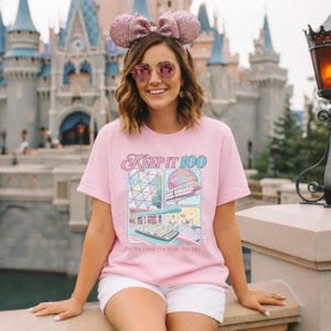 Op de afbeelding: Lichtroze T-shirt met de tekst "Keep It 100" en kleurrijke illustraties van een themapark. Het shirt heeft ook de tekst "On The Land, The Seas, The Sky".