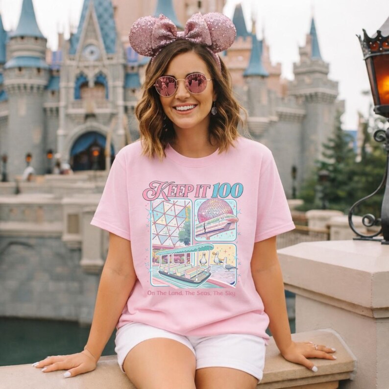 Op de afbeelding: Lichtroze T-shirt met de tekst "Keep It 100" en kleurrijke illustraties van een themapark. Het shirt heeft ook de tekst "On The Land, The Seas, The Sky".