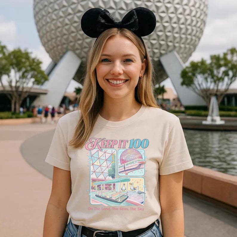 Op de afbeelding: Beige t-shirt met de tekst "Keep It 100" en kleurrijke illustraties van een themapark. Het shirt wordt gedragen met zwarte muisoren. De achtergrond toont een grote, bolvormige structuur en bomen.