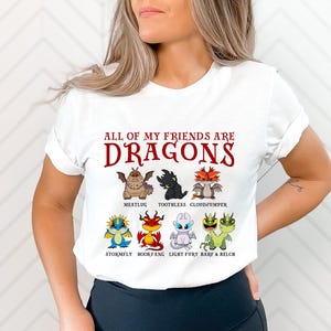 May include: White t-shirt with the text "All of my friends are dragons" in red. The shirt features a cartoon illustration of eight dragons, including Toothless, Meatlug, Cloudjumper, Stormfly, Bookfang, Light Fury, Barf, and Belch.