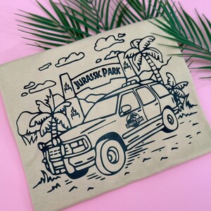 May include: A brown t-shirt with a black line drawing of a Jeep driving through a jungle with palm trees and the words "Jurassic Park" on a sign.